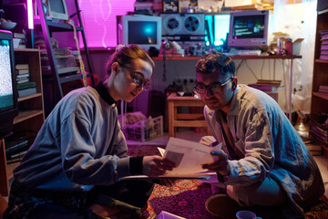 Two excited diverse young conspiracy theorists studying criminal profiles while sitting on carpet in dark messy room