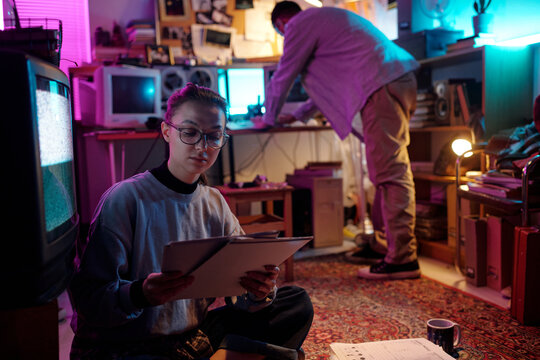 Young woman holding white folder with documents while sitting in messy room, her unknown male colleague using computer in background