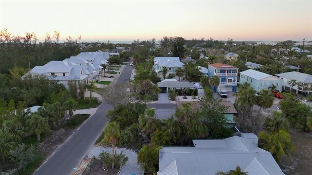 Florida neighborhood with coastal architecture,white roofs designed to reflect sunlight. Elevated designs help mitigate flooding risks,while lush landscaping and palm trees enhance. Aerial rising shot