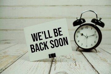 We'll Be Back Soon text on paper card with alarm clock on wooden background