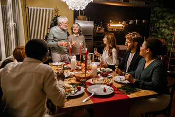 An elderly White couple in their 70s, standing at the head of a table in a room that is decorated for Christmas, and giving a toast as their whole family has dinner together, in a dimly-lit room