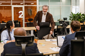 A middle-aged White businessman in his 50s, smiling as he looks over a table full of his younger...