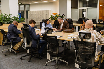 A middle-aged White businessman in his late 50s, helping his mid-30s Asian female colleague with some papers as they sit among their coworkers at a large table in an office hall
