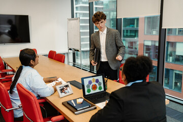 A young White man in his late 20s, talking to his older Black female superior as he stands up from the table in an office meeting room, with a young Indian female colleague sitting across from him