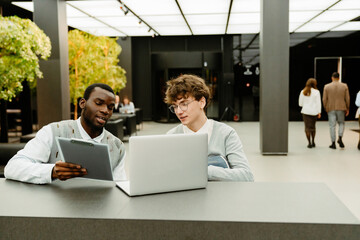 A young White man in his early 20s, having a conversation with his older Black male colleague, showing him his laptop, as they sit together in a spacious office hall, and the man shows him a clipboard