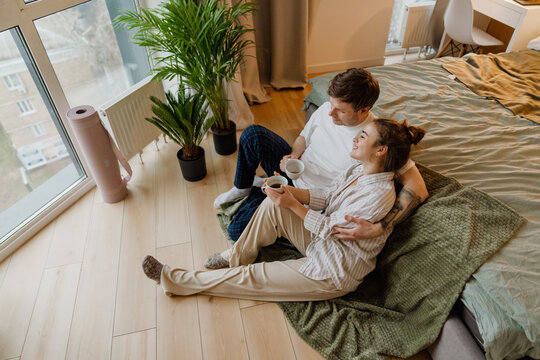 A happy young Caucasian couple relaxes on the floor near a bed in their home and smiles. The man embraces the woman holding a cup. Potted plants and large windows are captured in the room. - Powered by Adobe