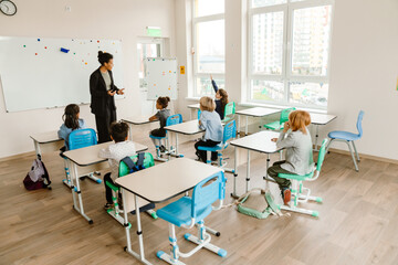 A Black female teacher in her 30s standing in a bright classroom with big windows and looking at a White school-aged girl sitting at a desk with a raised hand next to a multiethnic group of classmates