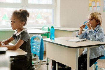 Obraz premium A young, school-aged White boy with short blond hair, sitting at his desk in a classroom and listening, with a pencil in his hand, as his Black female classmate sits in front of him