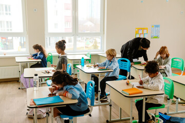 A Black female teacher in her 30s, who is dressed in a black business suit, standing next to the desk of one of her first-grade students, and leaning down to look the White boy in the face