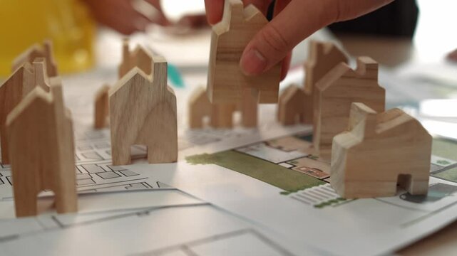 Architect and engineer discuss about building blueprint on table with architectural document, measurement tools scatter around at office, use wooden block for house positioning. Closeup. Delineation.