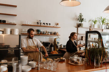 An adult White male employee in a white apron works behind the counter, while a young White woman in a black outfit prepares coffee in a cafe. Wooden shelves and kitchen equipment fill the space.
