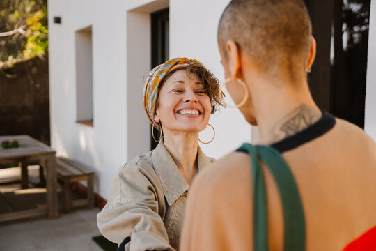 An adult White smiling woman with curly hair, a patterned headband, and large hoop earrings interacts with a tattooed adult Caucasian woman. They converse in an outdoor area near a building. - Powered by Adobe