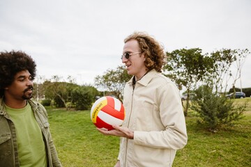 A young White man in his late 20s, with long, curly brown hair, holding a volleyball in his hand...