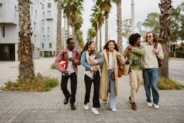 Fototapeta premium A young Asian woman in her late 20s, taking a stroll on a city street alongside a diverse group of her four young friends while holding her dachshund dog in her hands, on a sunny day