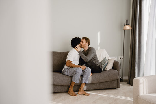A wholesome gay couple in their 30s, a Black man, and a White man, sitting together on a gray couch in a minimalistic white living room and kissing gingerly, as they're dressed in pajama clothes - Powered by Adobe