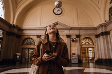 A young, brunette White woman in her late 20s, holding a phone in her hands and looking up in amazement as she stands inside of a spacious, beautiful train station