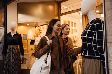 A young brunette White woman in her late 20s, looking at a sweater that is displayed on a mannequin on the street, as her young blond White female friend stands beside her, as they're near a shop