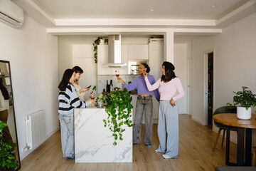 Fototapeta premium Four diverse women gather in a modern well-lit kitchen and celebrate. One holds a glass of wine while the marble-topped island holds potted plants and adds to the stylish design of the space.