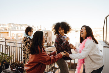 A young Latina woman in her late 20s, dancing around on the balcony of a high-rise apartment as her three young female friends laugh and have fun, on a sunny day, with a view of a town in the back