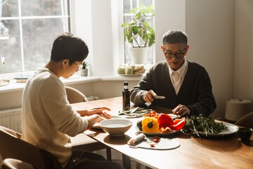An elderly Asian grandpa in his 70s, cutting some vegetables together with his adult grandson, who is in his 30s, and having a conversation, as they sit at the table in a living room