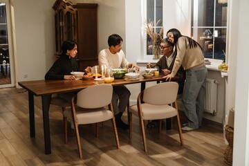 Fototapeta premium A happy Asian family of four shares a meal at a wooden dining table. The parents and a grandson engage in conversation with an elderly grandfather while eating from a variety of dishes and drinks.