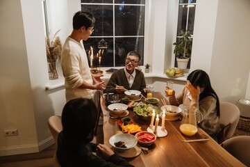 An Asian family talks sitting and standing at a table full of food, and has dinner, at the end of the table sits a senior man who talks to a man in his 30s who stands at the table, in a dining area.
