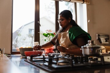 An Indian woman in her 30s, wearing an apron over a green t-shirt, salting a bowl of food while standing next to the stove in a modern kitchen, with the window in the background lighting her