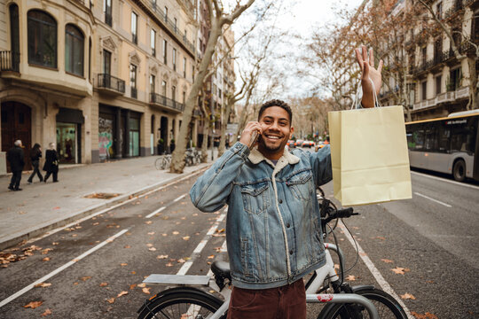 A Black man in his mid-20s, dressed in a denim jacket, waving to someone while talking on the phone as he holds a gift bag in his hand, standing in the bicycle lane on the road of a city