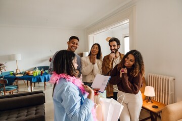 In a sunlit living room, four multiethnic friends in their late 20s congratulate their Black female friend in her 20s on her birthday, as she reads a birthday card and holds gifts in shopping bags.