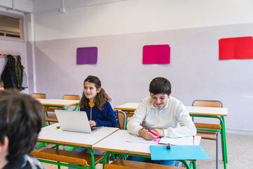 Girl using laptop and boy writing on notebook studying and learning in elementary school classroom