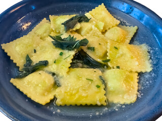 Square ravioli pasta in a light sauce with sage leaves are served in a blue ceramic bowl. The dish...