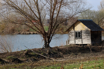 The photograph depicts a small wooden hut located on the shore of a body of water. The hut has a...