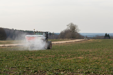Fototapeta premium Tractor spraying crops in a rural field during early spring with a scenic background and cloudy sky