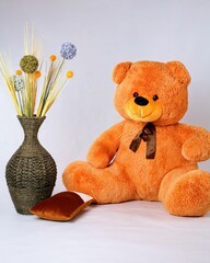 On a white background, there is a beautiful big orange bear, and next to it is a vase with flowers in the shape of a sphere.