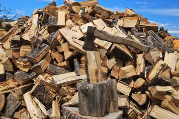 Axe Embedded in a Log with a Large Pile of Firewood in the Background. A rustic axe stuck in a wooden chopping block, surrounded by a large pile of freshly split firewood. The rough texture of the log