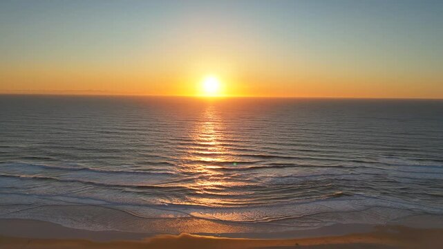 Drone captures golden sunset reflecting across textured ocean waves with foamy surf rolling onto a quiet sandy shoreline in soft, peaceful light at the edge of day.