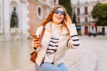 A stylish woman walks around a sunny city with a phone. A beautiful tourist in sunglasses is...