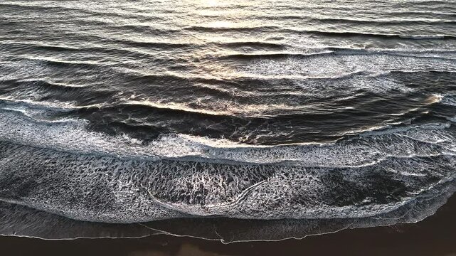 Aerial view of textured ocean waves breaking in layers on a dark wet beach, with soft reflections and foamy surf at sunset, captured in a peaceful and atmospheric moment by drone. 