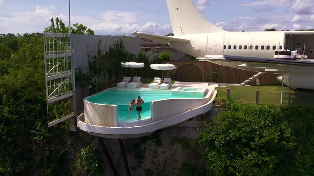 Couple relaxing in infinity pool on their luxury exotic hotel made from airplane. Aircraft repurposed for private villa in Bali. VIP vacation in luxurious setting of unique design. 