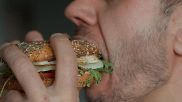 Man takes big bite of seeded bagel filled with egg, cheese, tomato and greens. Hungry man eats sandwich at kitchen, close up