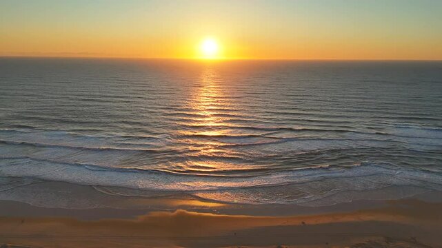 Drone captures a top-down view of golden sunset light reflecting on ocean surf, with textured waves rolling gently onto a peaceful sandy shoreline in a calm and tranquil seascape. 