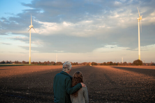 Grandfather and granddaughter standing in a field with wind turbines at sunset