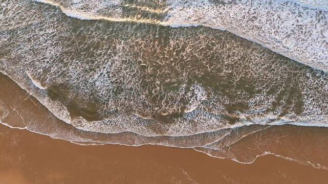 Aerial view of ocean surf patterns from above, with foamy waves washing over a golden sandy beach in calm, warm light at sunset a peaceful and textured coastal moment. 