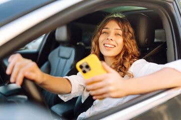 Young woman with curly hair with a phone in her hands driving a car. Happy female driver with a...