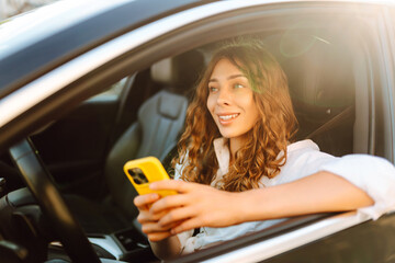 Young woman with curly hair with a phone in her hands driving a car. Happy female driver with a...