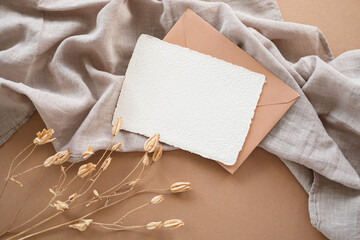 Close-up overhead view of a blank White deckle edge card and envelope on a table with a decorative dried plant and linen fabric on a table