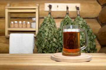 In the sauna interior a glass mug of light beer on a wooden tray stands on a plank bench on the background of a log wall with dry birch brooms and shelf with bottles of aromatic oils and white towel. 