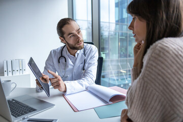 Male doctor sitting at desk in hospital holding tablet discussing digital health consultation