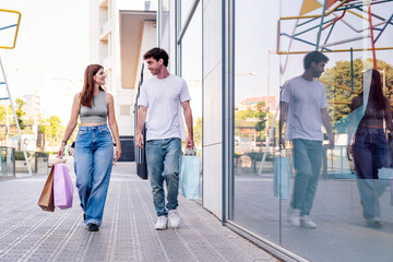 young adult man and a woman in casual clothes walking down a sidewalk near of a mall carrying shopping bags, concept of couple and urban lifestyle, copyspace for text