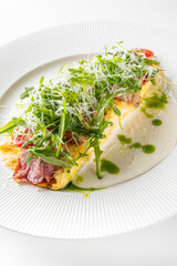 A beautifully plated omelette filled with fresh greens, tomatoes, and sliced meat, drizzled with herb sauce on a white background.